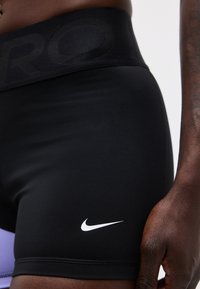 Μαύρο αθλητικό σορτς με λευκό λογότυπο Nike στο μηρό, φαρδιά μέση και ορατό μέρος του ισχίου και του χεριού ενός ατόμου.