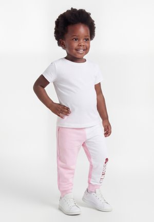 Niño pequeño sonriente con peinado afro, de pie con la mano en la cadera, vistiendo camiseta blanca, pantalones deportivos rosas y blancos, y zapatillas blancas sobre fondo liso.