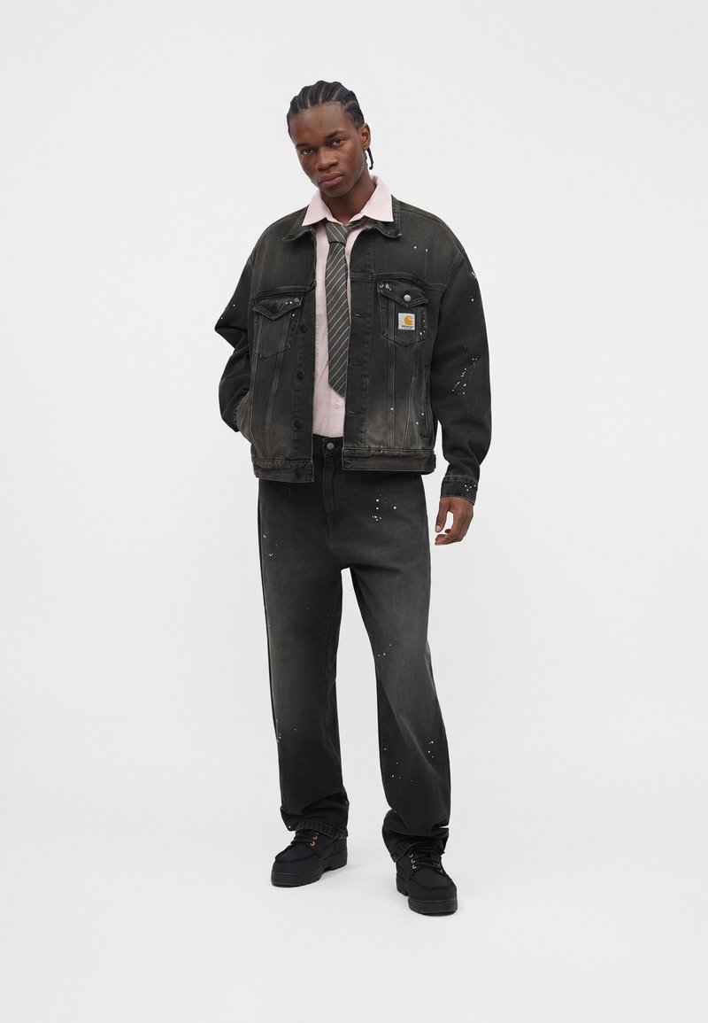 Man with braided hair wearing black denim jacket and pants with paint splatters, light pink shirt, striped tie, and black shoes standing against white background.