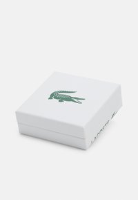 Hvid firkantet boks med tekstureret overflade, med et grønt krokodillelogo på toppen og "LACOSTE" trykt på siden.