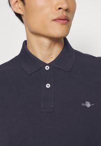 Marineblå polo skjorte laget av teksturert bomull med tre-knappslukking, med hvite knapper og en liten brodert logo på brystet.