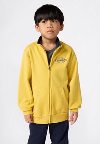 Sudadera amarilla con cremallera hecha de tela suave, con cuello azul, logo en el pecho y bolsillos frontales. Usada sobre una camiseta gris.