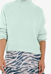 Helles mintgrünes Sweatshirt mit hohem Kragen, geripptem Muster und übergroßen Ärmeln, kombiniert mit einem gemusterten Rock in Blau und Beige.