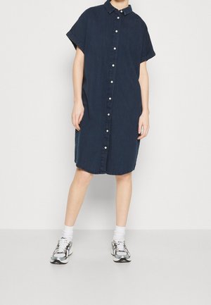 Person trägt ein lockeres, dunkelblaues, kurzärmeliges Hemdkleid mit weißen Knöpfen, weiße Socken und silberne und schwarze Sneakers.