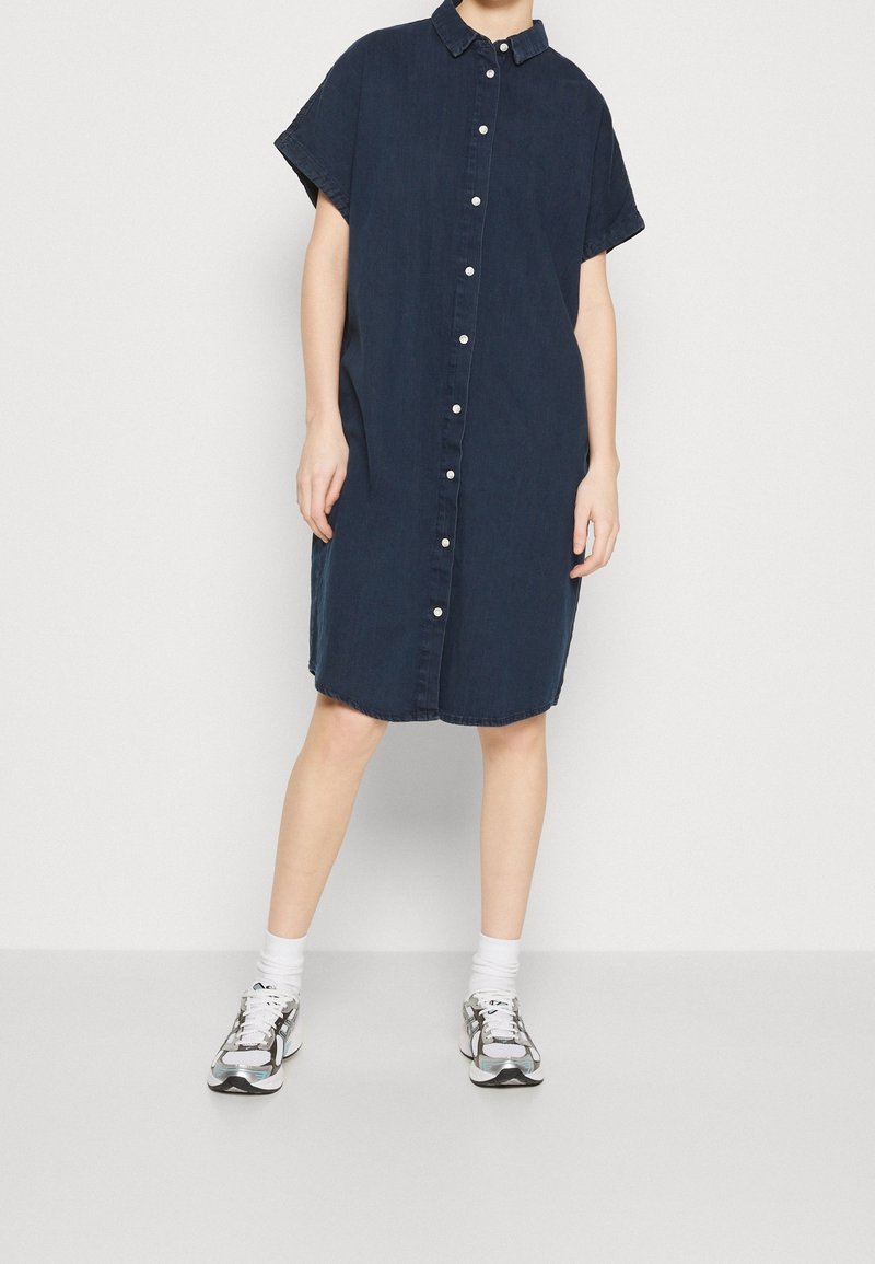 Personne portant une robe ample bleu foncé à manches courtes boutonnée avec des boutons blancs, des chaussettes blanches et des baskets argentées et noires.