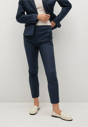 Kobieta poprawiająca granatowe, dopasowane spodnie noszone z pasującą marynarką, kremowym topem i kremowymi mokasynami na jednolitym jasnym tle.