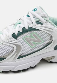 Białe i turkusowe buty sportowe z siateczkowego materiału, z srebrnym akcentem oraz teksturowanym, wzorzystym logo "N" po boku.