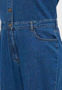 Combinaison en denim bleu foncé, avec un devant boutonné, une taille ceinturée et des poches avant. Le tissu a une finition lisse et légèrement texturée.