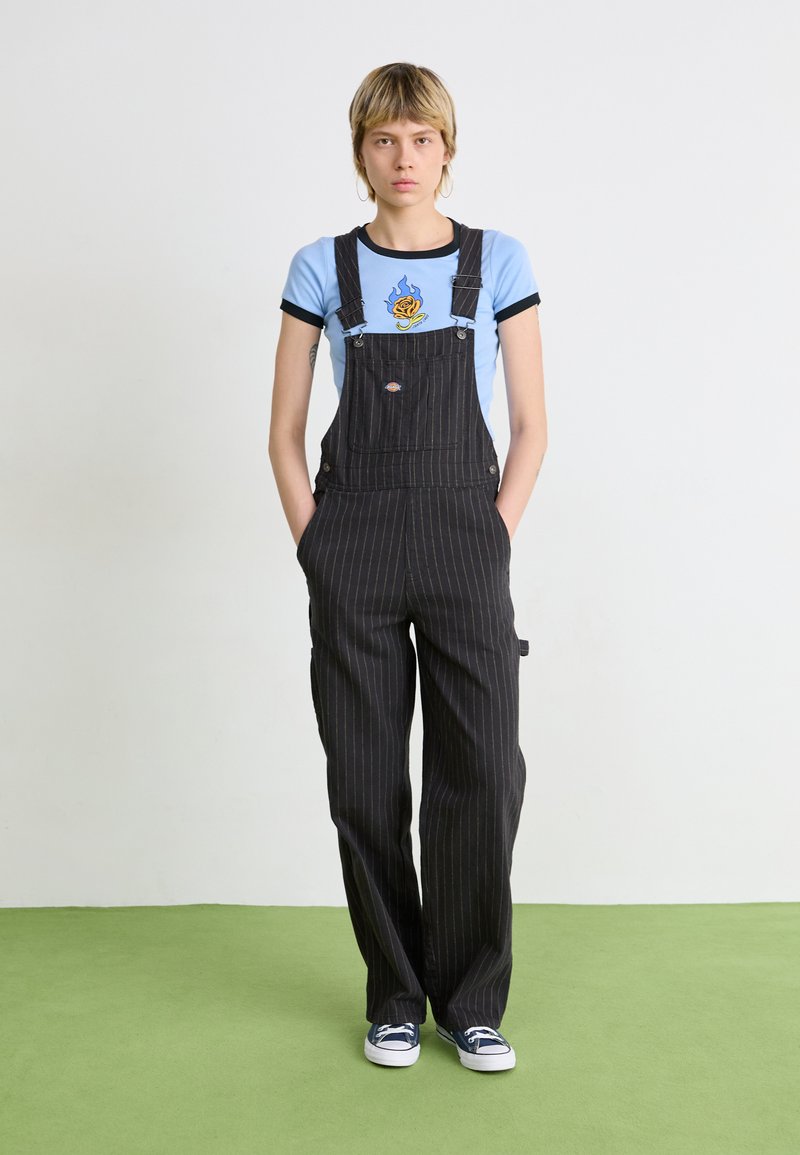 Schwarze Latzhose mit Nadelstreifen und einer Fronttasche, kombiniert mit einem hellblauen T-Shirt mit Flammenmuster und dunkelblauen Sneakern.