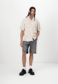 Kortermet beige skjorte med hvit abstrakt mønster, sammen med grå denimshorts. Svarte klumpete sko og hvite sokker fullfører antrekket.