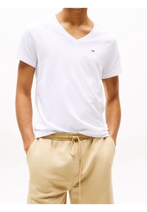 Mann trägt weißes V-Ausschnitt-T-Shirt mit kleinem Logo und beige Kordelzug-Shorts, Hände in den Taschen, steht vor weißem Hintergrund.