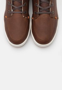 Bruna läderskor med en slät yta, rund tå och kontrasterande vit gummisula. Har svarta snören och orange sytt accent.