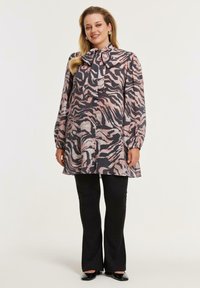 Kvinde iført en langærmet bluse med tigermønster og en stor sløjfe om halsen, sorte flagrende bukser og sorte spidse sko, smilende.
