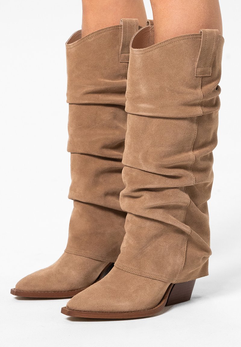 Beige suède laarzen met een slouch-stijl, scherpe neuzen en lage blokhakken. Ze hebben een zachte textuur en een gathered ontwerp dat tot halverwege de kuit reikt.