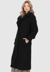 Cappotto lungo in pile nero con colletto, tasche laterali e texture morbida, mostrato su uno sfondo grigio chiaro.