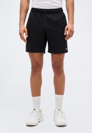 Mann in schwarzen Sportshorts, weißen Sportsocken, weißen Turnschuhen und weißem Hemd, der vor einem schlichten Hintergrund steht.