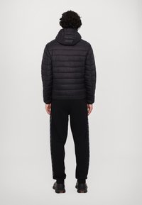 Sort polstret jakke med hætte, der har en quilted tekstur. Bæres sammen med sorte sweatpants med et subtilt sidelæns mønster og sorte sko.