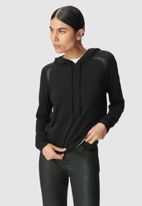 Mujer con cabello liso lleva un suéter negro con capucha y pantalones negros, sujetando el dobladillo del suéter, mirando hacia su izquierda.