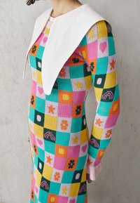 Robe tricotée colorée avec un large col blanc, présentant un motif en patchwork de carrés en rose, teal, jaune et noir, avec divers motifs.