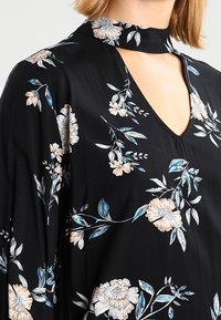 Schwarzes Blumenkleid mit V-Ausschnitt und Schlüsselloch-Ausschnitt, langen Ärmeln und einem weichen Stoff, der mit pinken und blauen Blumenmustern verziert ist.