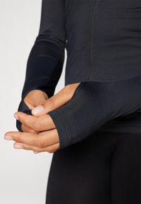 Fekete, hosszú ujjú sportfelső, hüvelykujjnak kialakított nyílásokkal, texturált anyagból, testhezálló fazonnal, a test előtt összekulcsolt kezekkel.