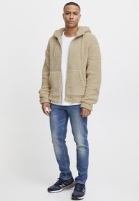 Beige fleece zip-up hettegenser med en teksturert overflate, to frontlommer og snorhette, kombinert med en hvit t-skjorte og blå jeans.