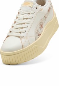 Valkoinen nahkainen alustakengät vaaleanruskeilla yksityiskohdilla, tekstuuripohjalla ja kukkakirjontayksityiskohdilla sivuilla. Varustettu valkoisilla nauhoilla.