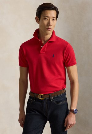 Homme portant un polo rouge avec le col relevé, un jean foncé, une ceinture marron et une montre-bracelet avec un cadran bleu et un bracelet en cuir marron.
