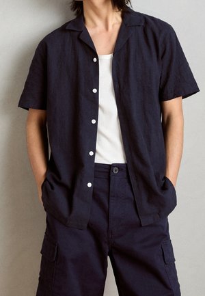 Osoba ubrana w biały tank top pod granatową koszulę z krótkim rękawem zapinaną na guziki oraz granatowe spodnie typu cargo, ręce w kieszeniach na jednolitym tle.