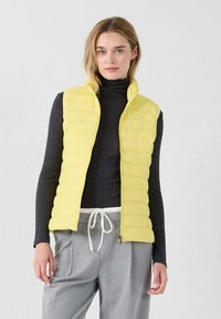 Chaleco acolchado amarillo ligero con un diseño de acolchado vertical, cuello alto y un logotipo sutil. Lleva puesto sobre un suéter de cuello alto oscuro y pantalones grises.