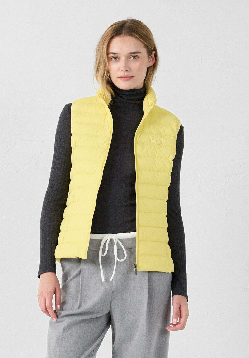 Chaleco acolchado amarillo ligero con un diseño de acolchado vertical, cuello alto y un logotipo sutil. Lleva puesto sobre un suéter de cuello alto oscuro y pantalones grises.