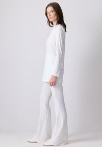 Donna in piedi di lato che indossa una tunica bianca a maniche lunghe e pantaloni a palazzo a coste su uno sfondo bianco puro.