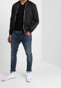 Černá bomber bunda s bílým zipem, nošena přes černé tričko s výstřihem do V. V kombinaci s modrými džíny a bílými teniskami, s ohrnutými rukávy.