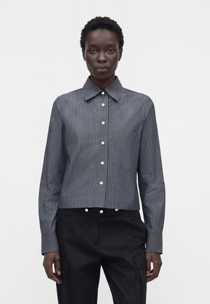 Femme portant une chemise gris foncé à fines rayures, boutonnée, à manches longues, et un pantalon noir, sur un fond blanc uni.