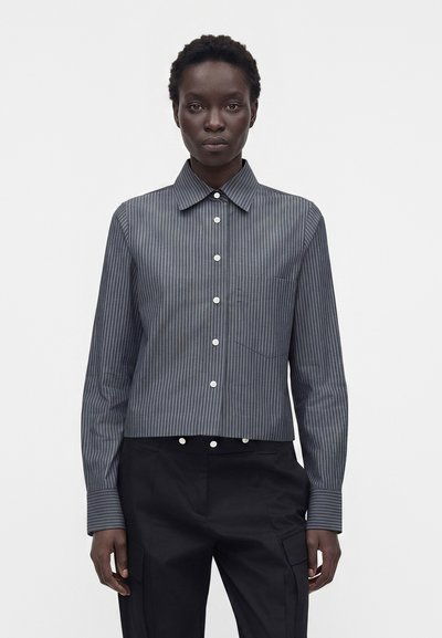 Femme portant une chemise gris foncé à fines rayures, boutonnée, à manches longues, et un pantalon noir, sur un fond blanc uni.