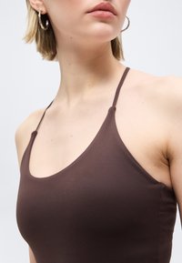 Κοντινό πλάνο ενός ατόμου που φοράει καφέ αμάνικο τοπ με λεπτές τιράντες και σκουλαρίκια κρίκους, εμφανίζοντας την περιοχή του λαιμού και των ώμων.