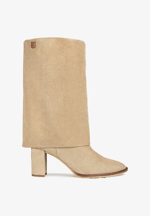 Botín de ante beige hasta el tobillo con un tacón bloques medio y un diseño de solapa. Presenta una punta afilada y un parche de marca cosido en la parte superior.