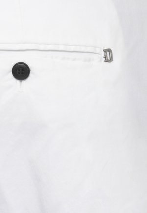 Tessuto bianco con un bottone nero a quattro fori e una piccola cerniera metallica vicino a una cucitura orizzontale o al bordo di una tasca.