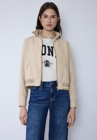 Veste bomber beige texturée avec poignets côtelés, fermeture à glissière et poches latérales, portée sur un t-shirt blanc graphique et un jean bleu.
