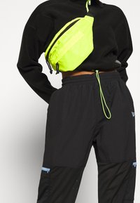 Sudadera negra, riñonera reflectante en un brillante amarillo neón, pantalones negros con detalles de malla y acentos de cremallera azules.