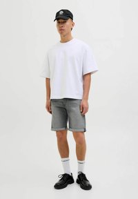 Witte T-shirt met korte mouwen, grijze denimshorts, zwarte schoenen en witte sokken met tekst. Zwarte pet met logo bovenop. Neutrale achtergrond.