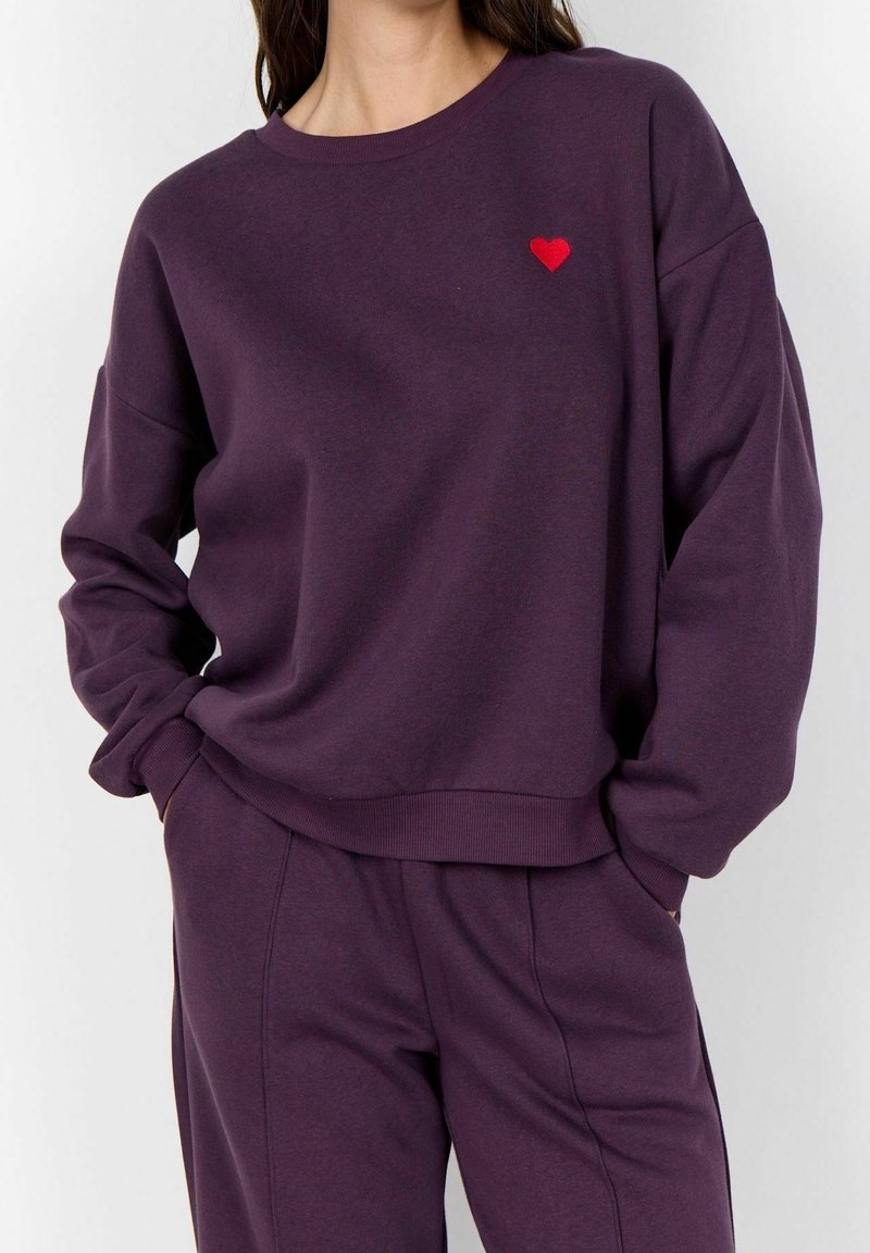 Femme portant un ensemble sweat-shirt et pantalon violet foncé avec un petit cœur rouge brodé sur la poitrine gauche, mains dans les poches du pantalon.