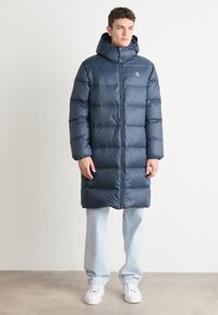 Marineblauer gepolsterter langer Mantel mit Kapuze, der einen Reißverschluss vorne hat und ein Logo auf der Brust trägt. Getragen über hellblauen Jeans und weißen Sneakers.
