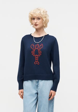 Mujer rubia con un suéter azul marino con diseño de langosta roja y vaqueros azul claro, complementado con un collar blanco de cuentas.