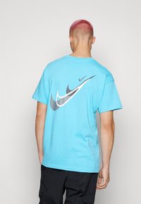 Persona con cabello corto rosa que lleva una camiseta Nike de color azul brillante con múltiples logotipos en blanco y negro en la parte posterior, pantalones negros.
