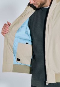 Beige Jacke mit hellblauem Innenfutter, ausgestattet mit einem Reißverschluss und einer Innentasche. Rippenstruktur auf der Außenseite.