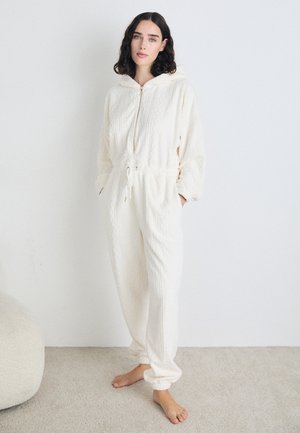 Krém színű, texturált anyagból készült overál, kapucnival, elöl cipzárral, húzózsinórral a deréknál, és elasztikus mandzsettákkal a ujjaknál és a bokán.
