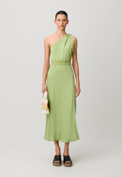 Donna che indossa un vestito midi monospalla verde chiaro, sandali neri con plateau, che tiene una piccola borsa beige intrecciata su uno sfondo semplice.