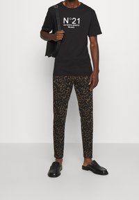 Czarna koszulka z białym napisem "N°21 VIA SANTO SPIRITO MILANO," w połączeniu z dopasowanymi brązowymi spodniami z ciemnym abstrakcyjnym wzorem i czarnymi butami wsuwanymi.