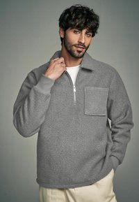 Harmaa fleece-pullovari, jossa on puolivetoketju etuosassa, rintatasku ja ribbauskahvat. Pehmeä rakenne ja rento muotoilu, yhdistettynä kermaisiin housuihin.